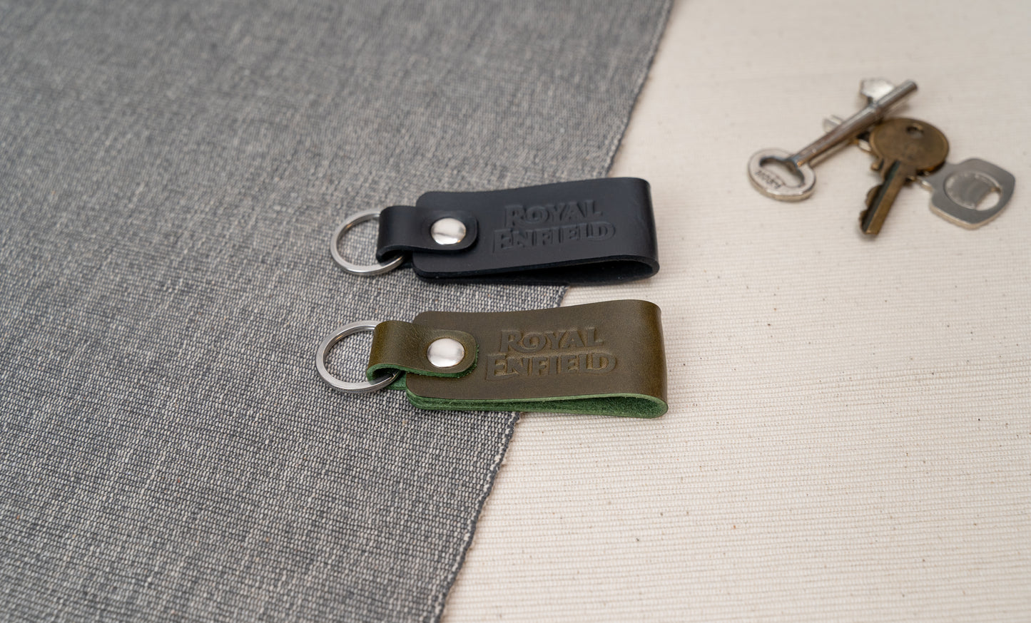 Two Royal Enfield leather keychains, one black and one olive green, on a textured surface with keys in the background.