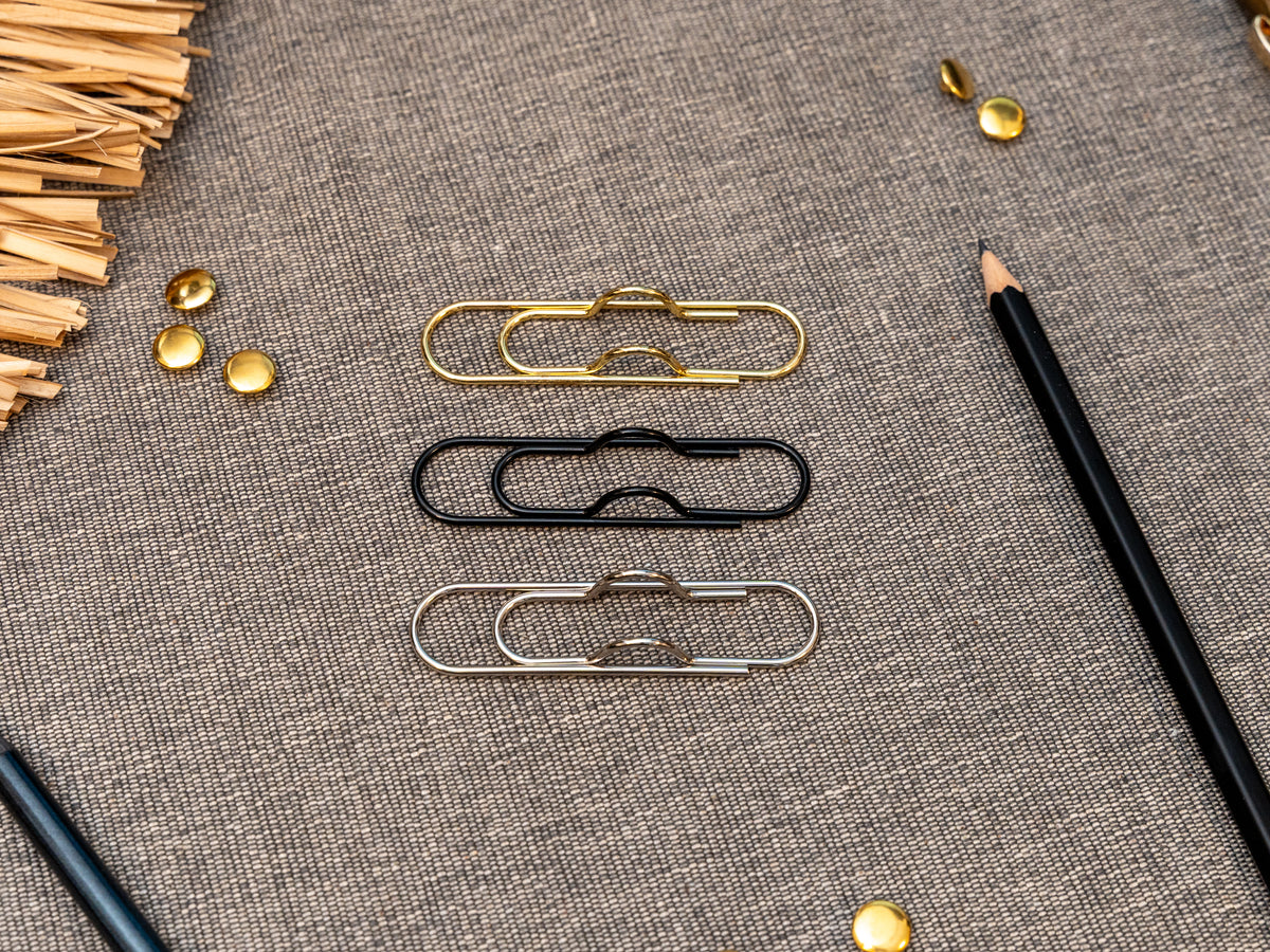 3 notebook pen holders in gold, black and silver that look like paperclips