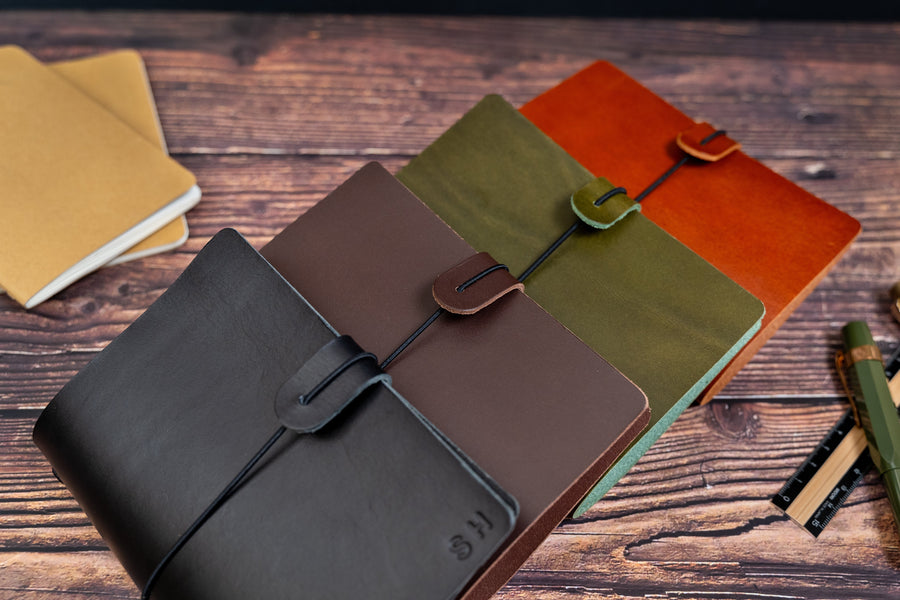 A collection of leather journal covers in various colors displayed on a wooden surface, with a pen and additional notebooks in the background.