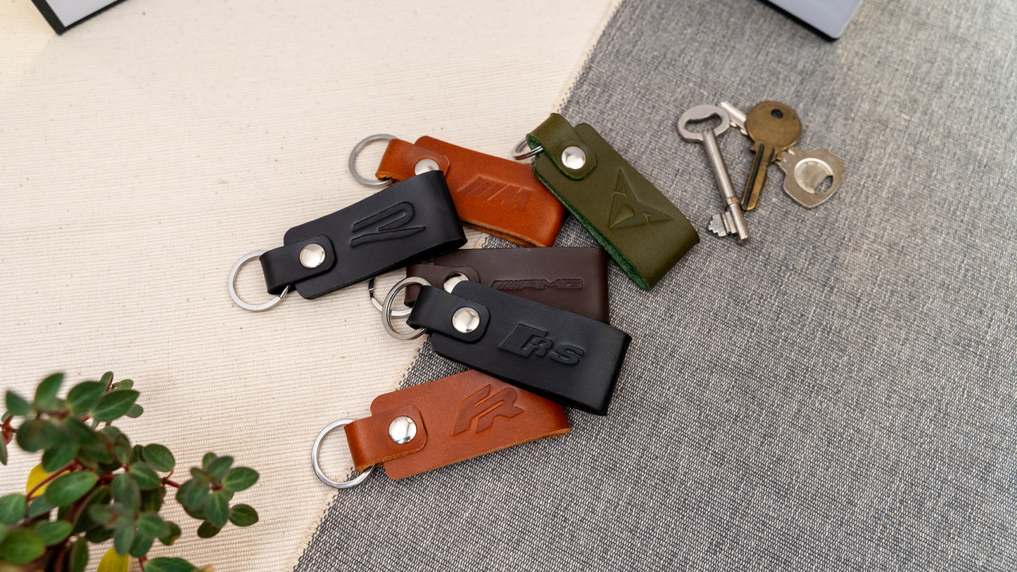 Mixture of keyrings in black, tan, olive green, and dark brown stamped with various logos such as Seat Cupra, VW R, BMW M Sport. Many car logos available in several colours with stainless steel or brass hardware