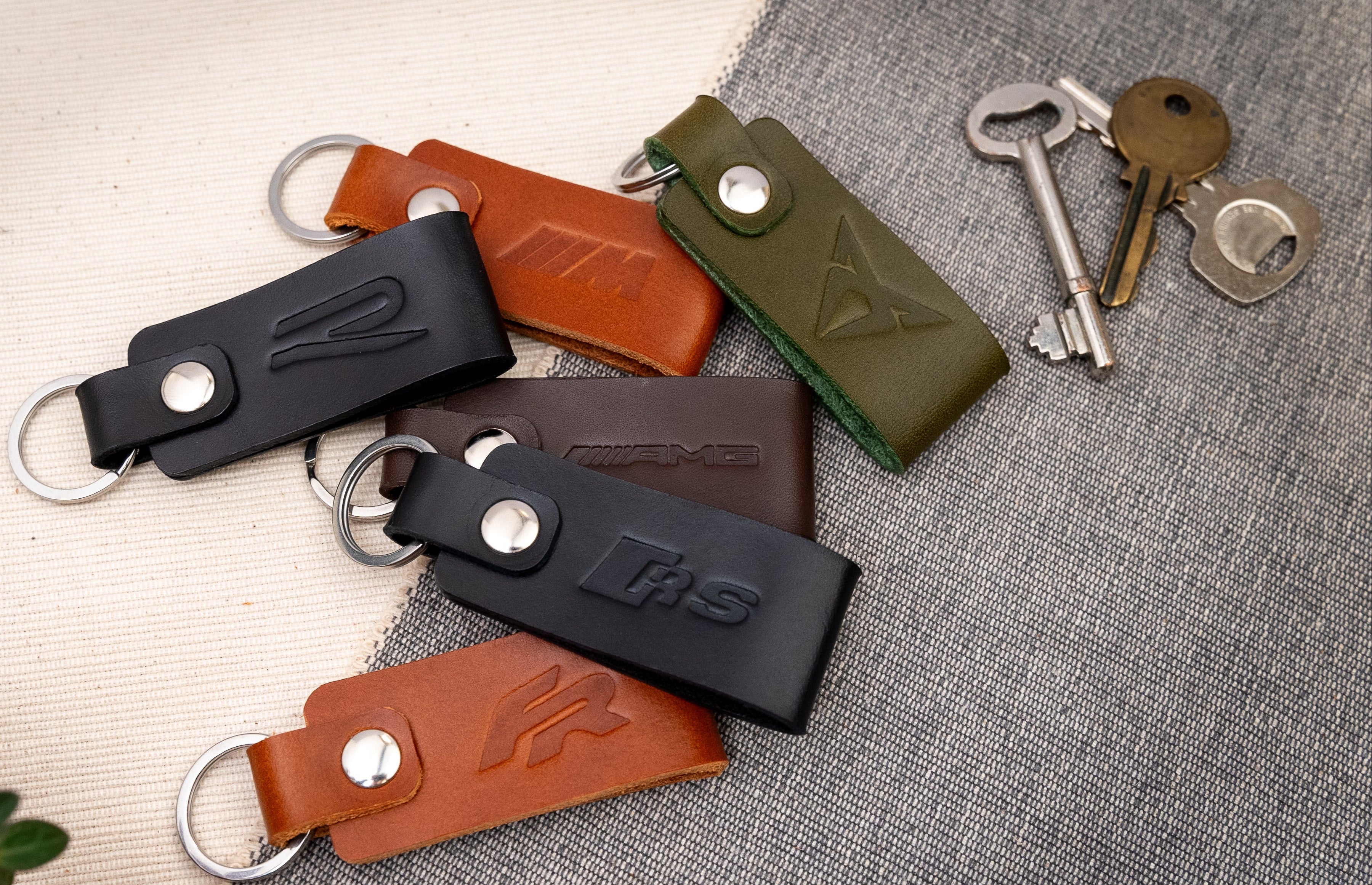 Collection of leather keychains debossed with car logos in black, brown, and olive green on a textured surface with keys.