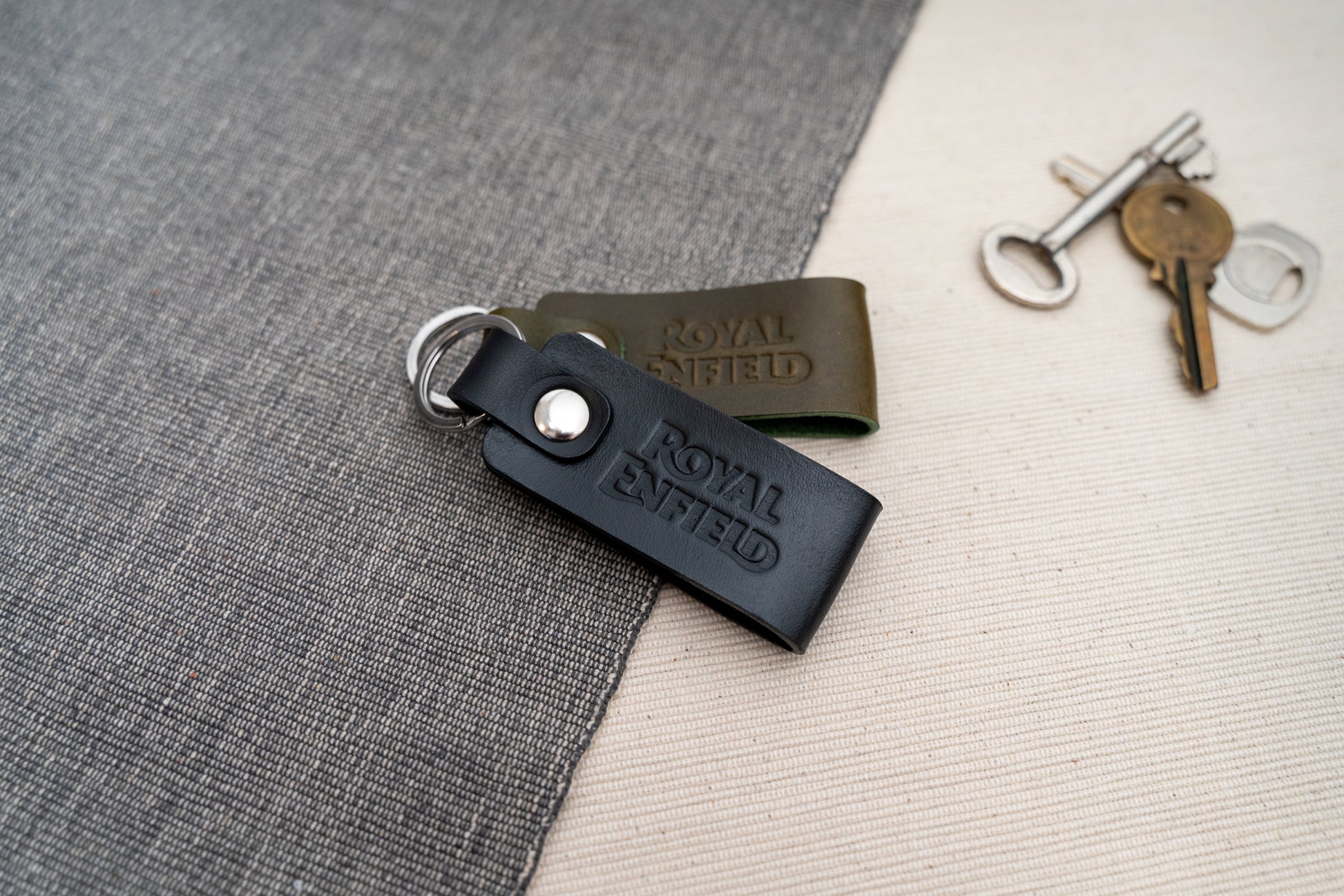 Two Royal Enfield leather keychains, one black and one olive green, on a textured surface with keys in the background.