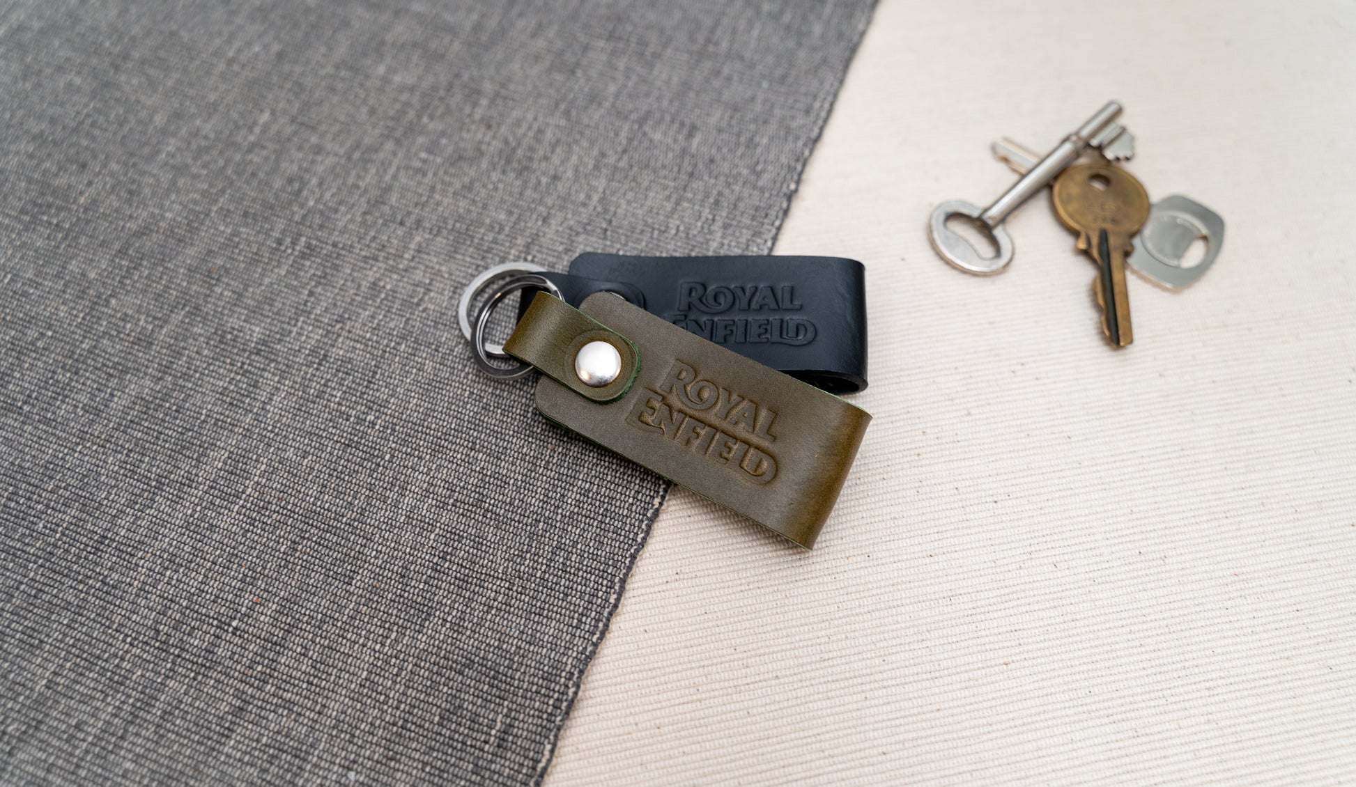 Two Royal Enfield leather keychains, one black and one olive green, on a textured surface with keys in the background.