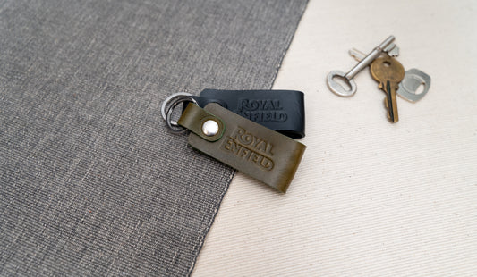 Two Royal Enfield leather keychains, one black and one olive green, on a textured surface with keys in the background.