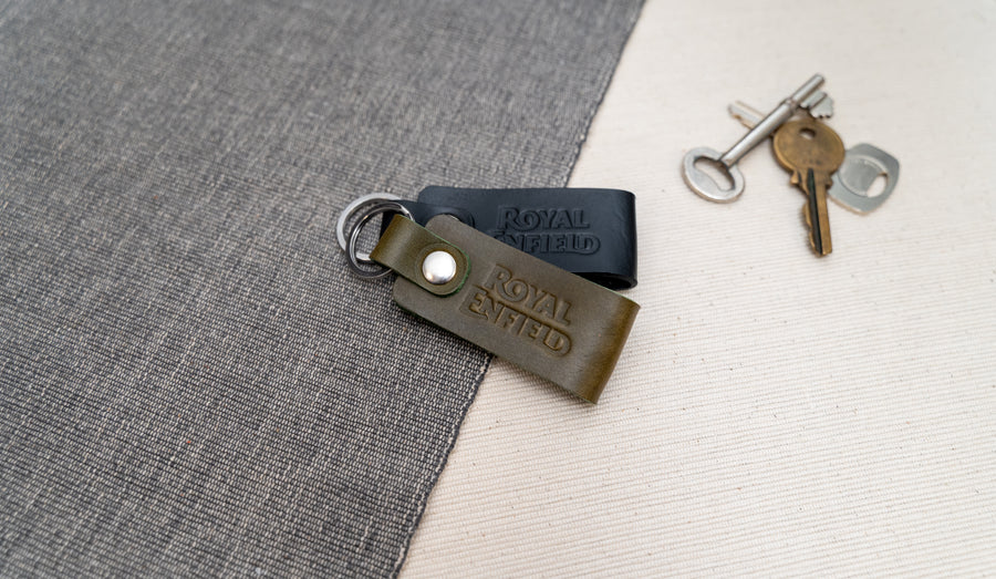 Two Royal Enfield leather keychains, one black and one olive green, on a textured surface with keys in the background.