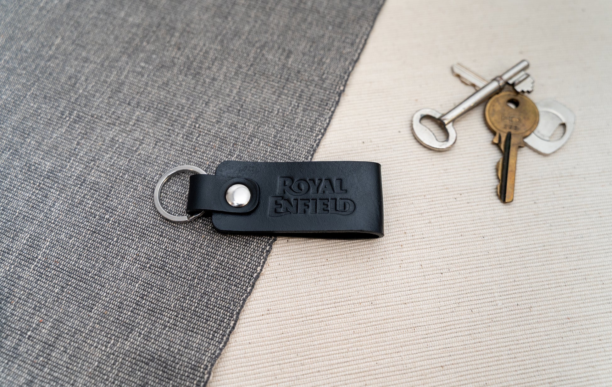 Black Royal Enfield leather keychain on a textured surface with keys in the background.