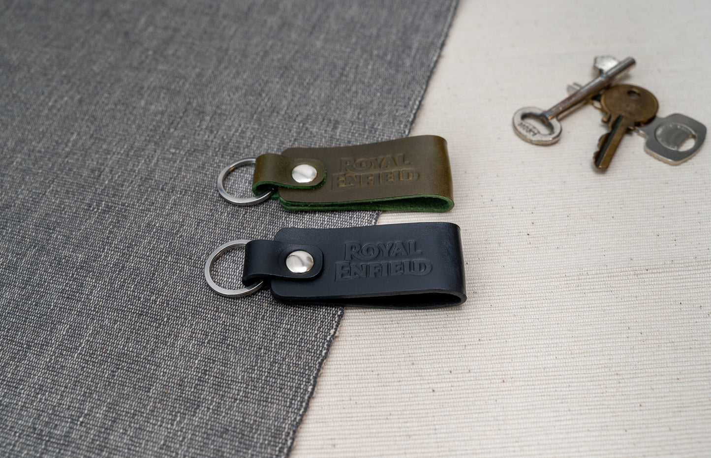 Two Royal Enfield leather keychains, one black and one olive green, on a textured surface with keys in the background.