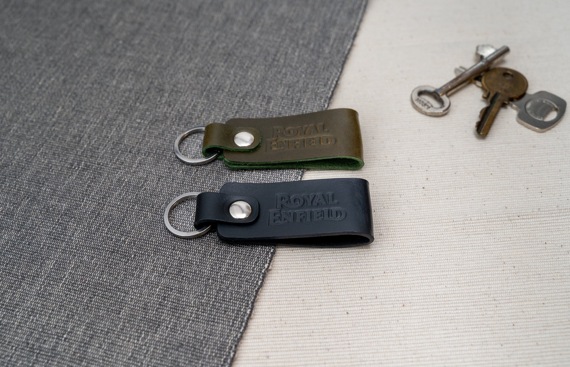 Two Royal Enfield leather keychains, one black and one olive green, on a textured surface with keys in the background.