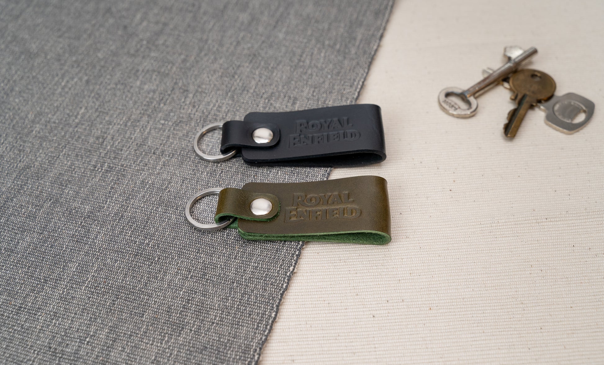 Two Royal Enfield leather keychains, one black and one olive green, on a textured surface with keys in the background.