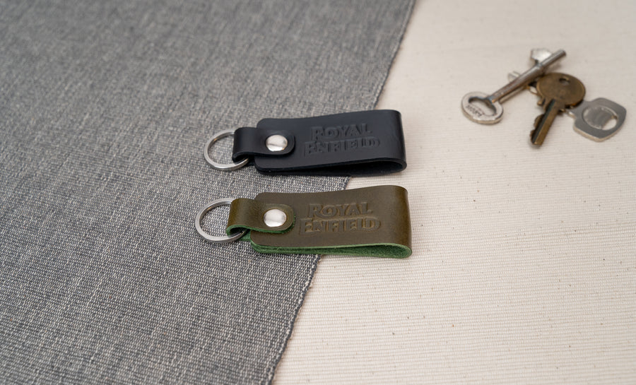Two Royal Enfield leather keychains, one black and one olive green, on a textured surface with keys in the background.