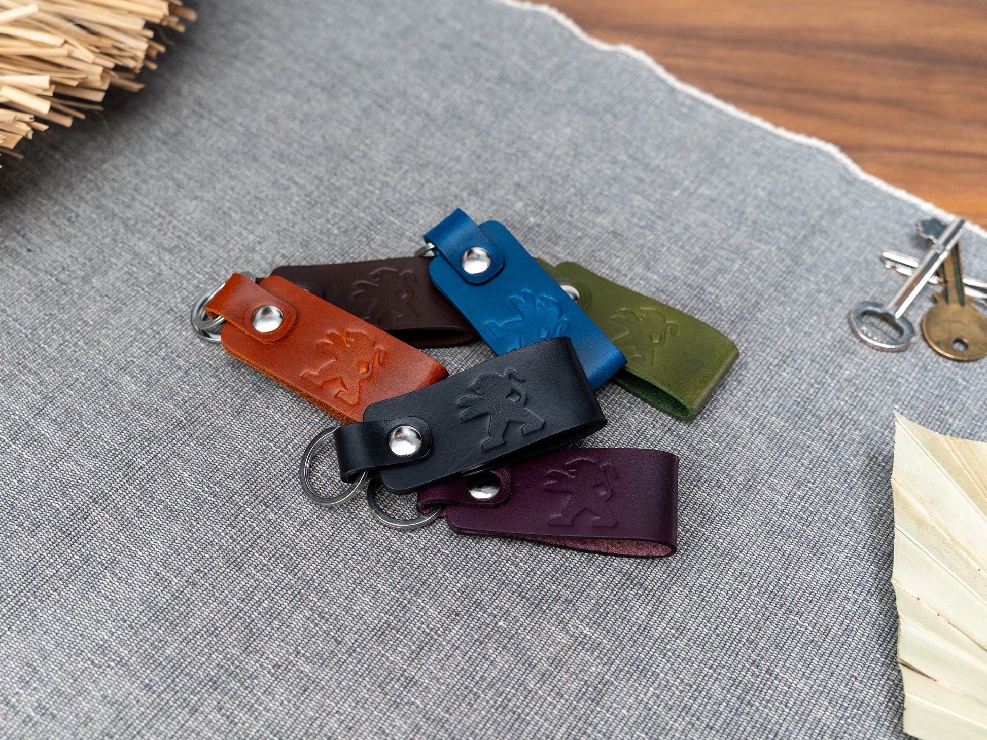 A group of folded leather keyrings in blue, tan, olive green, purple, dark brown and black leather with Peugeot car logo debossed on them. Made from Italian full grain leather