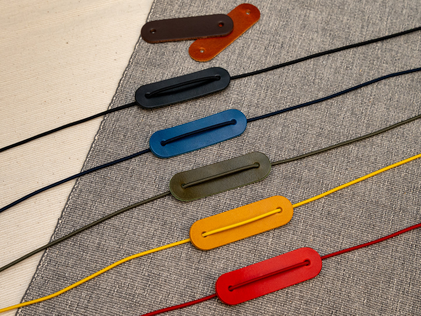 Six leather cord notebook closures in different colours (brown, tan, black, blue, olive green, yellow, and red) arranged diagonally on a textured grey fabric background.