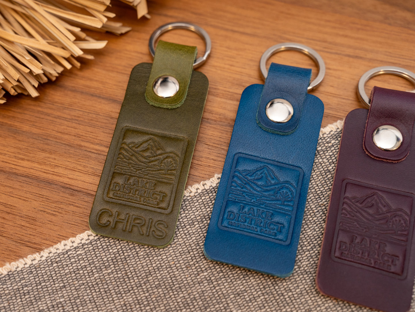 Several leather keyrings with a rectangular design in various colours such as olive green, navy blue and purple keyrings/keychains embossed with an image of the lake district. Stainless steel split ring and rivet.