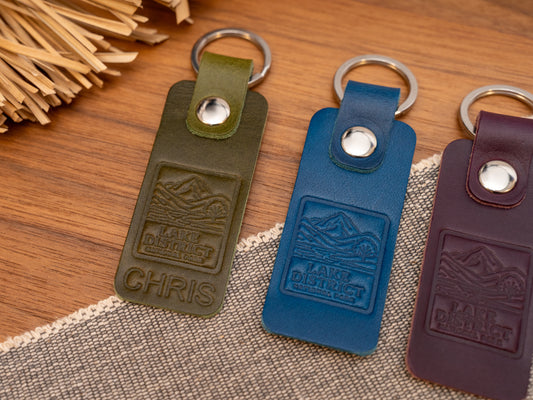 Several leather keyrings with a rectangular design in various colours such as olive green, navy blue and purple keyrings/keychains embossed with an image of the lake district. Stainless steel split ring and rivet.