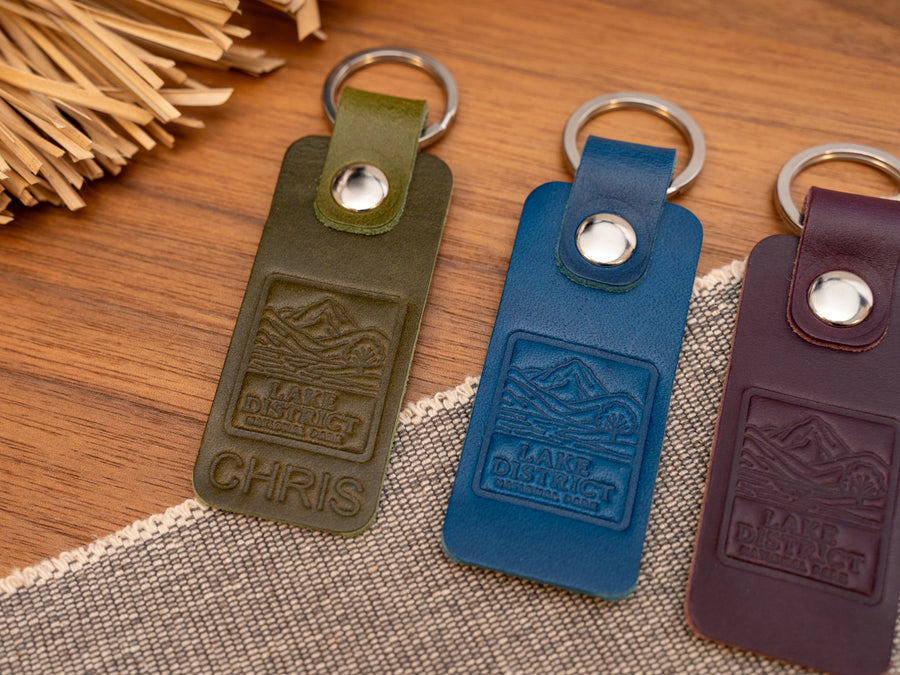 Several leather keyrings with a rectangular design in various colours such as olive green, navy blue and purple keyrings/keychains embossed with an image of the lake district. Stainless steel split ring and rivet.