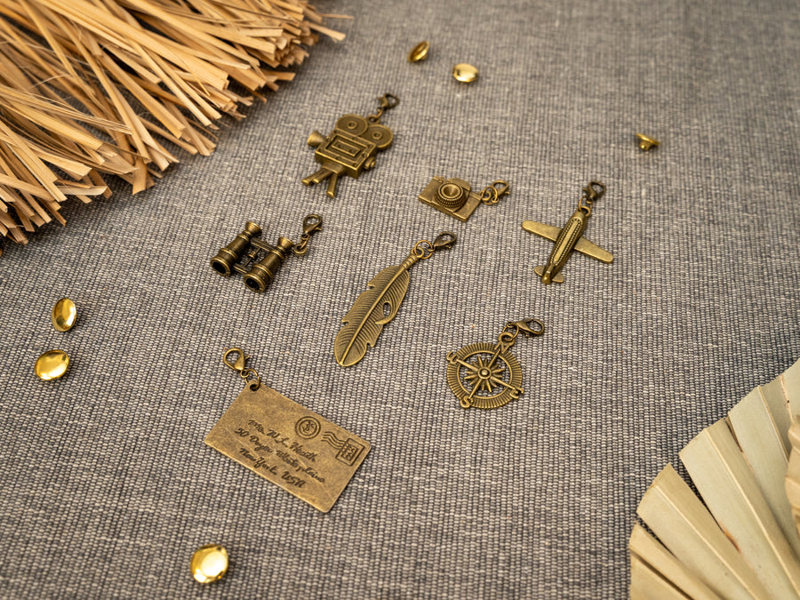 7 antique bass metal charms . The charms include a film camera, camera, a small airplane, binoculars, a feather, a compass, and an envelope.