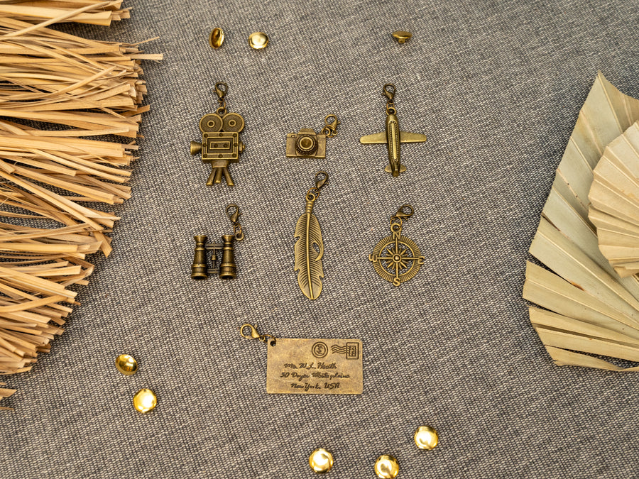 7 antique bass metal charms . The charms include a film camera, camera, a small airplane, binoculars, a feather, a compass, and an envelope.