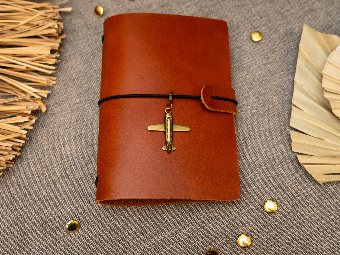 A tan leather journal with a black elastic band closure and a vintage brass aeroplane charm, displayed on a textured gray fabric background with decorative props.