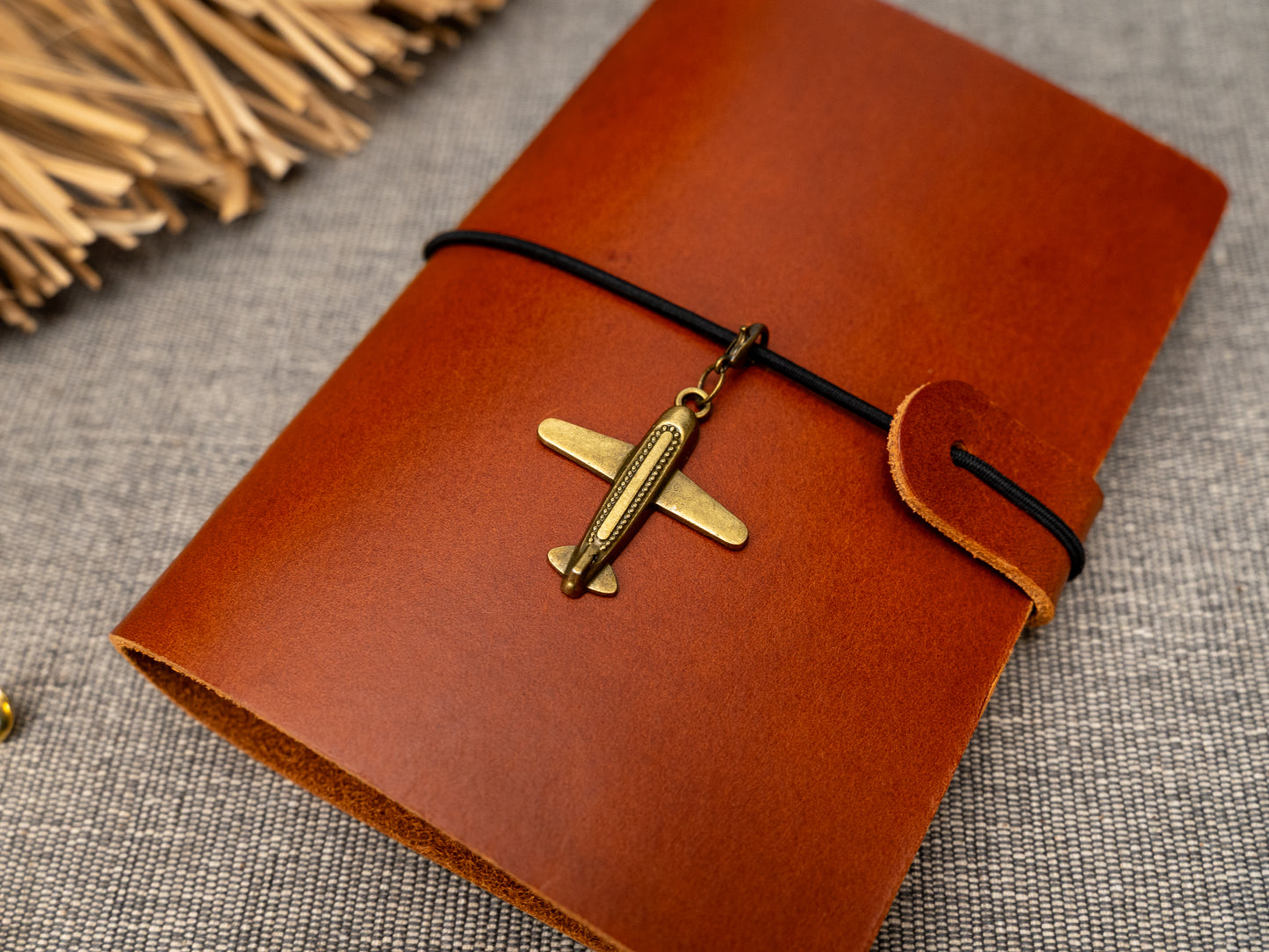 A tan leather journal with a black elastic band closure and a vintage brass aeroplane charm, displayed on a textured gray fabric background with decorative props.