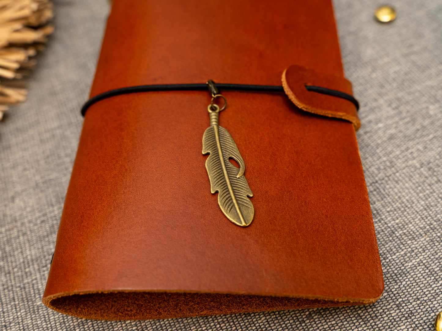 A tan leather journal with a black elastic band closure and a vintage brass feather charm, displayed on a textured gray fabric background with decorative props.