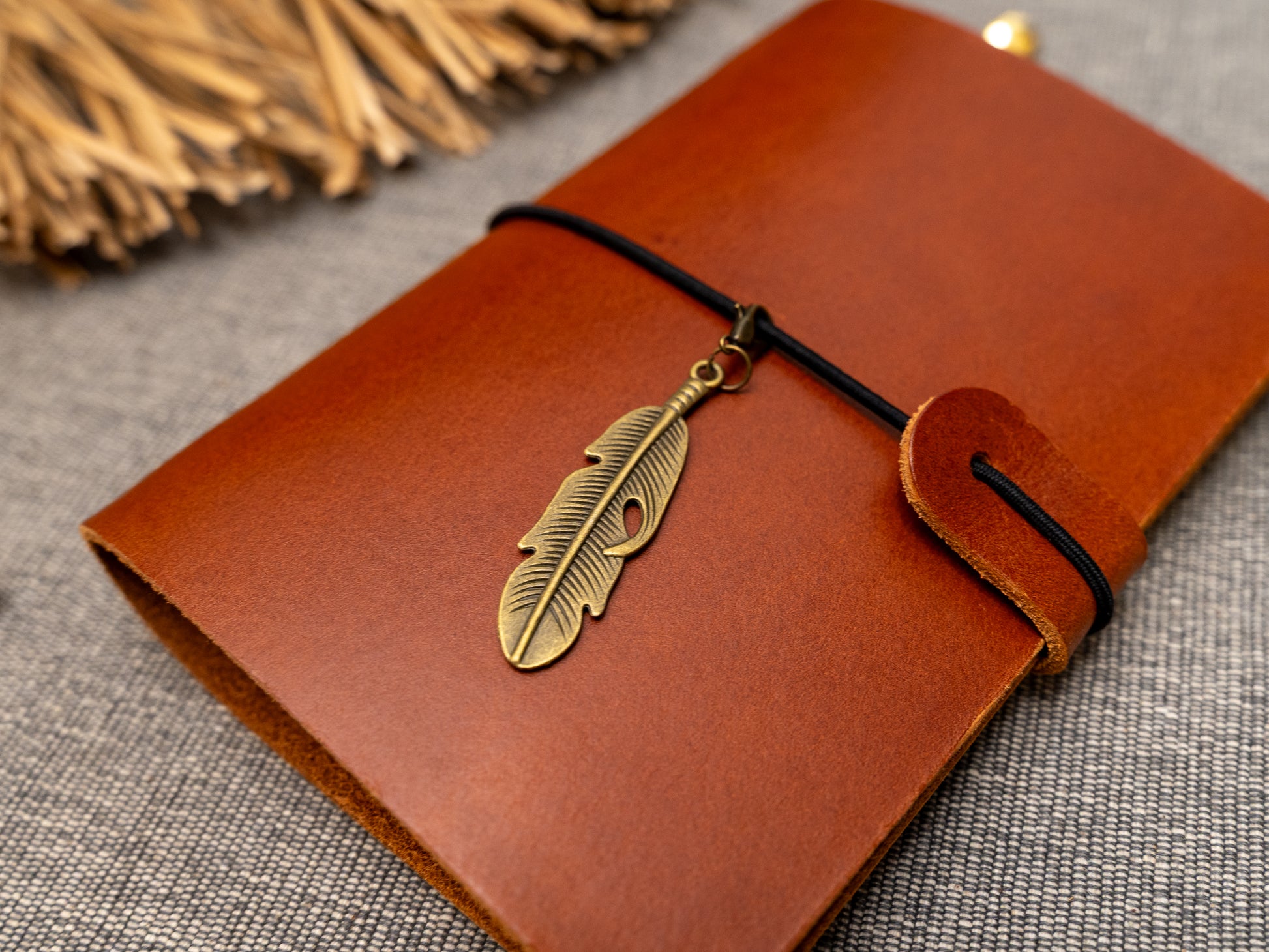 A tan leather journal with a black elastic band closure and a vintage brass feather charm, displayed on a textured gray fabric background with decorative props.