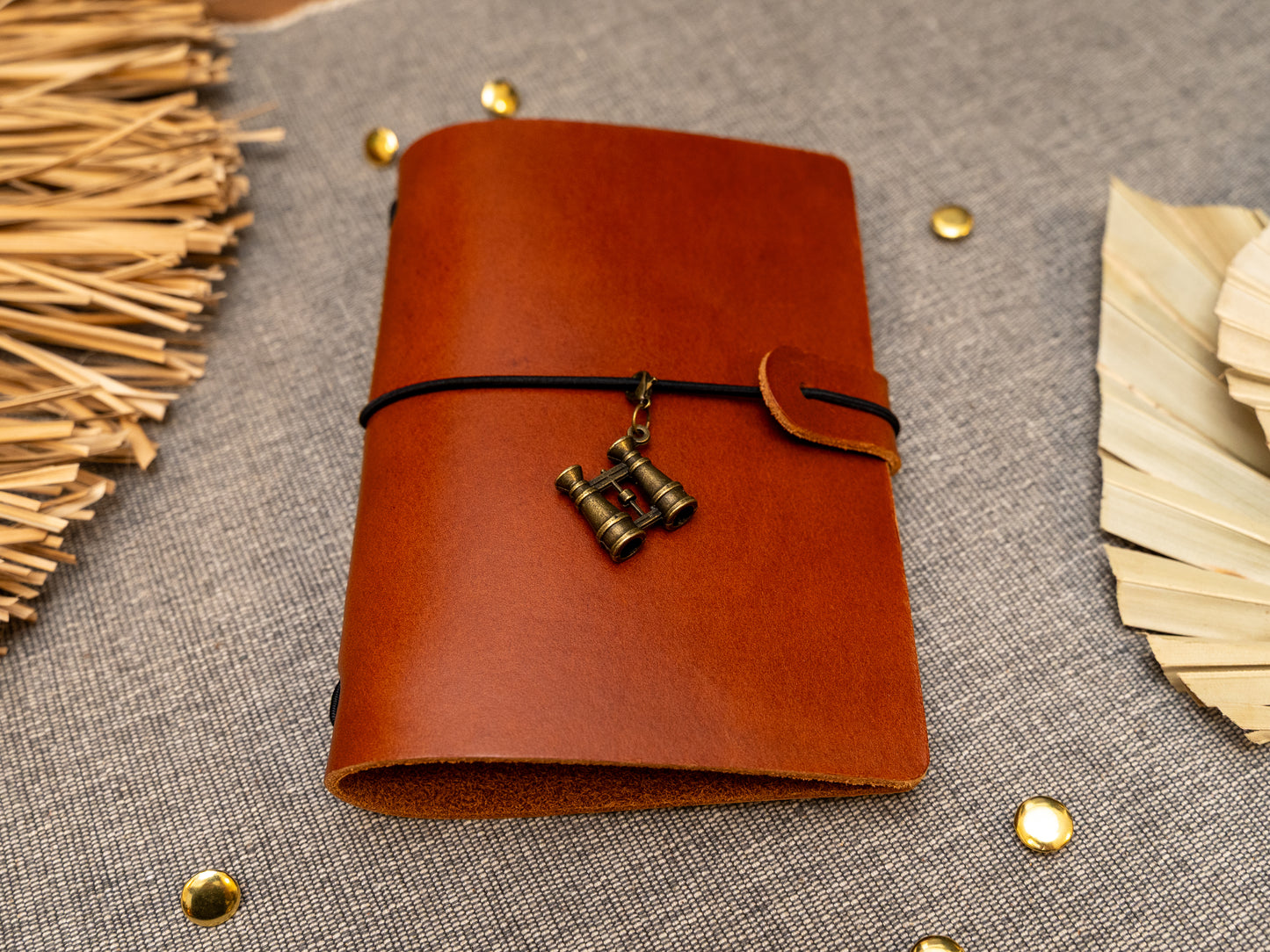 A tan leather journal with a black elastic band closure and a vintage brass binoculars charm, displayed on a textured gray fabric background with decorative props.