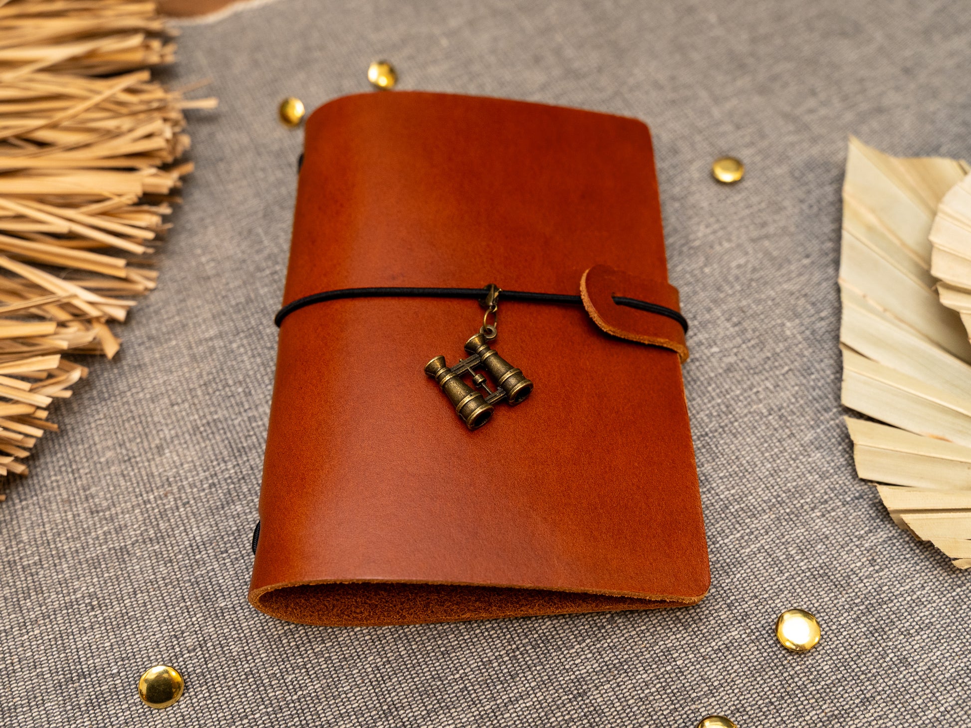 A tan leather journal with a black elastic band closure and a vintage brass binoculars charm, displayed on a textured gray fabric background with decorative props.