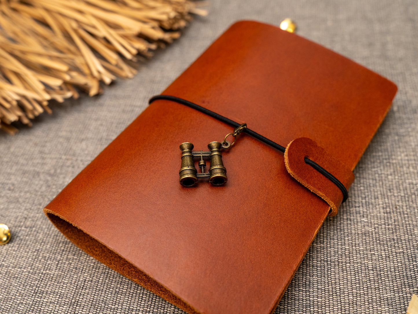 A tan leather journal with a black elastic band closure and a vintage brass binoculars charm, displayed on a textured gray fabric background with decorative props.