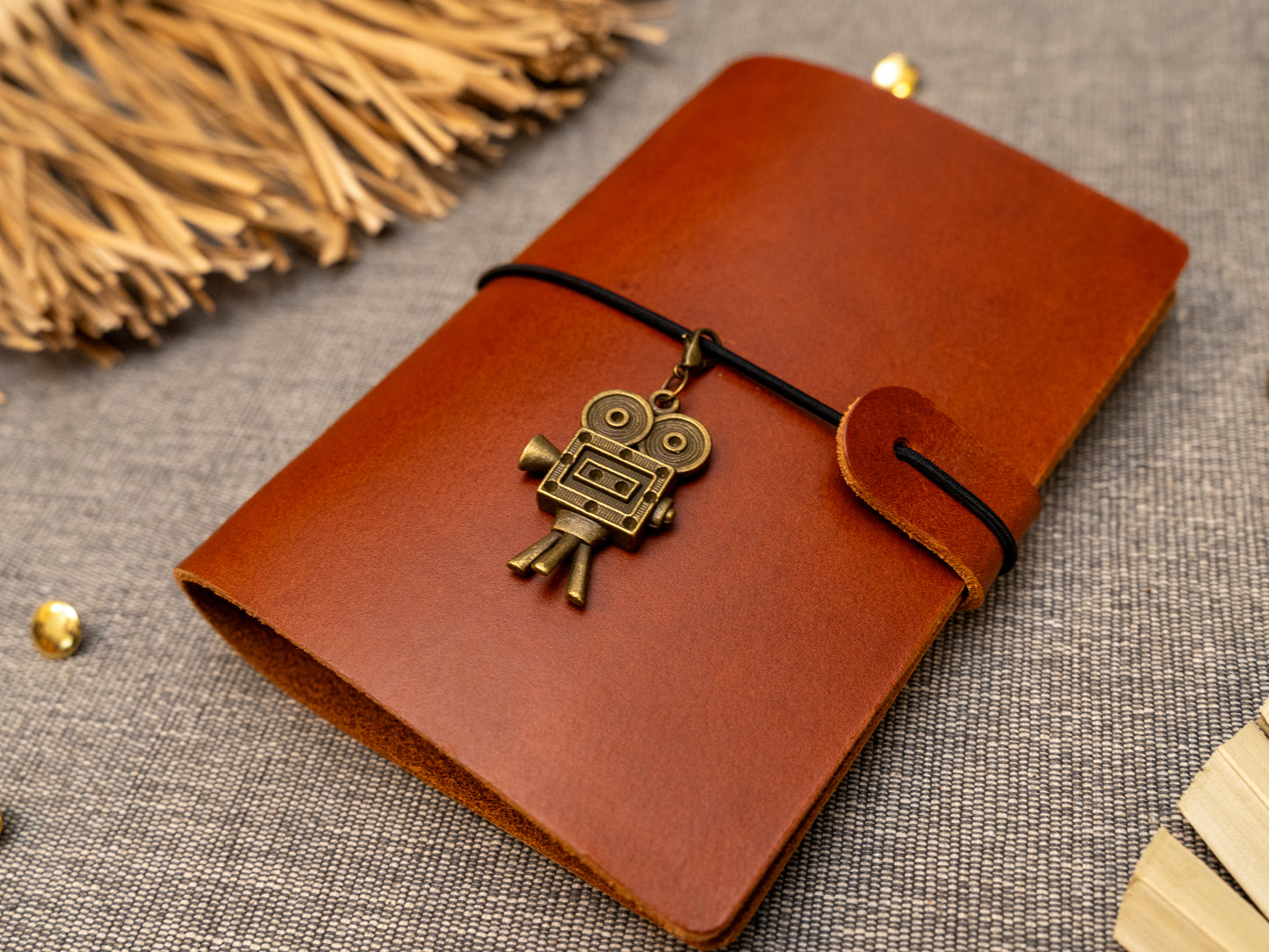 A tan leather journal with a black elastic band closure and a vintage film camera charm, displayed on a textured gray fabric background with decorative props.