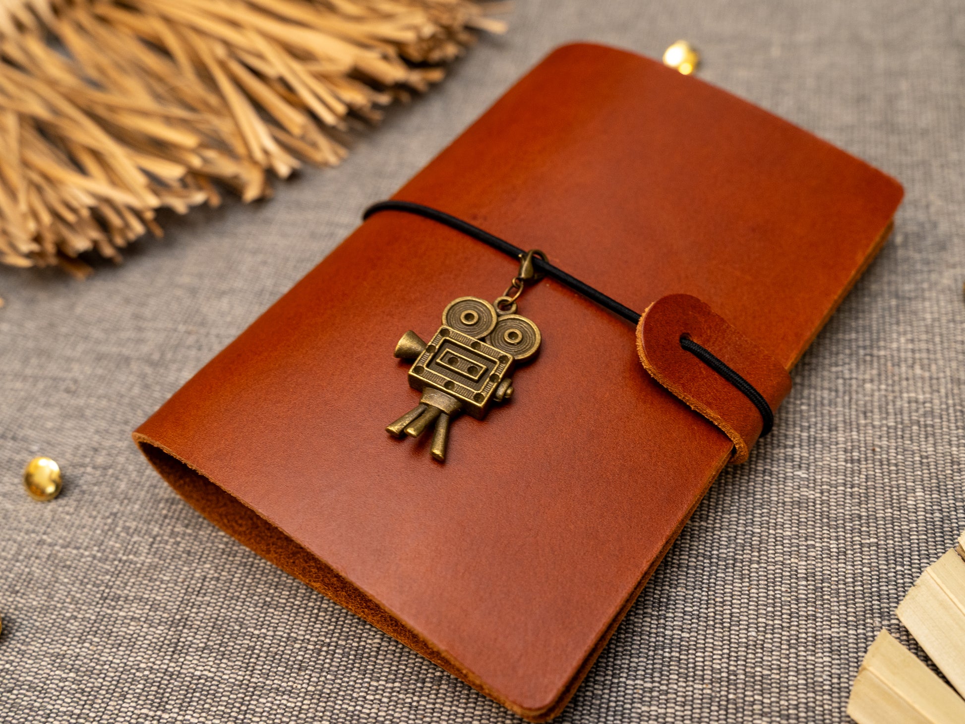 A tan leather journal with a black elastic band closure and a vintage film camera charm, displayed on a textured gray fabric background with decorative props.