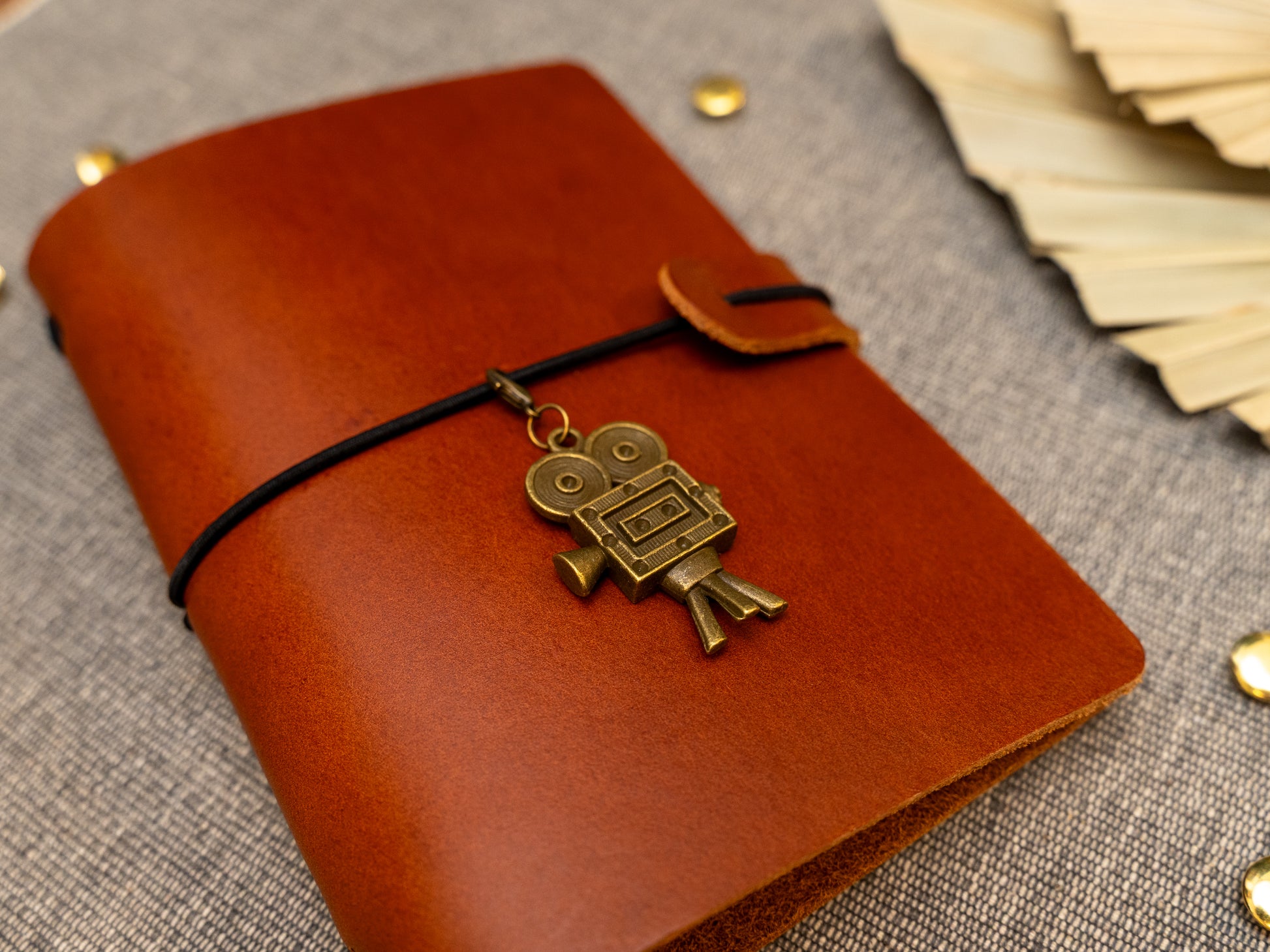 A tan leather journal with a black elastic band closure and a vintage film camera charm, displayed on a textured gray fabric background with decorative props.