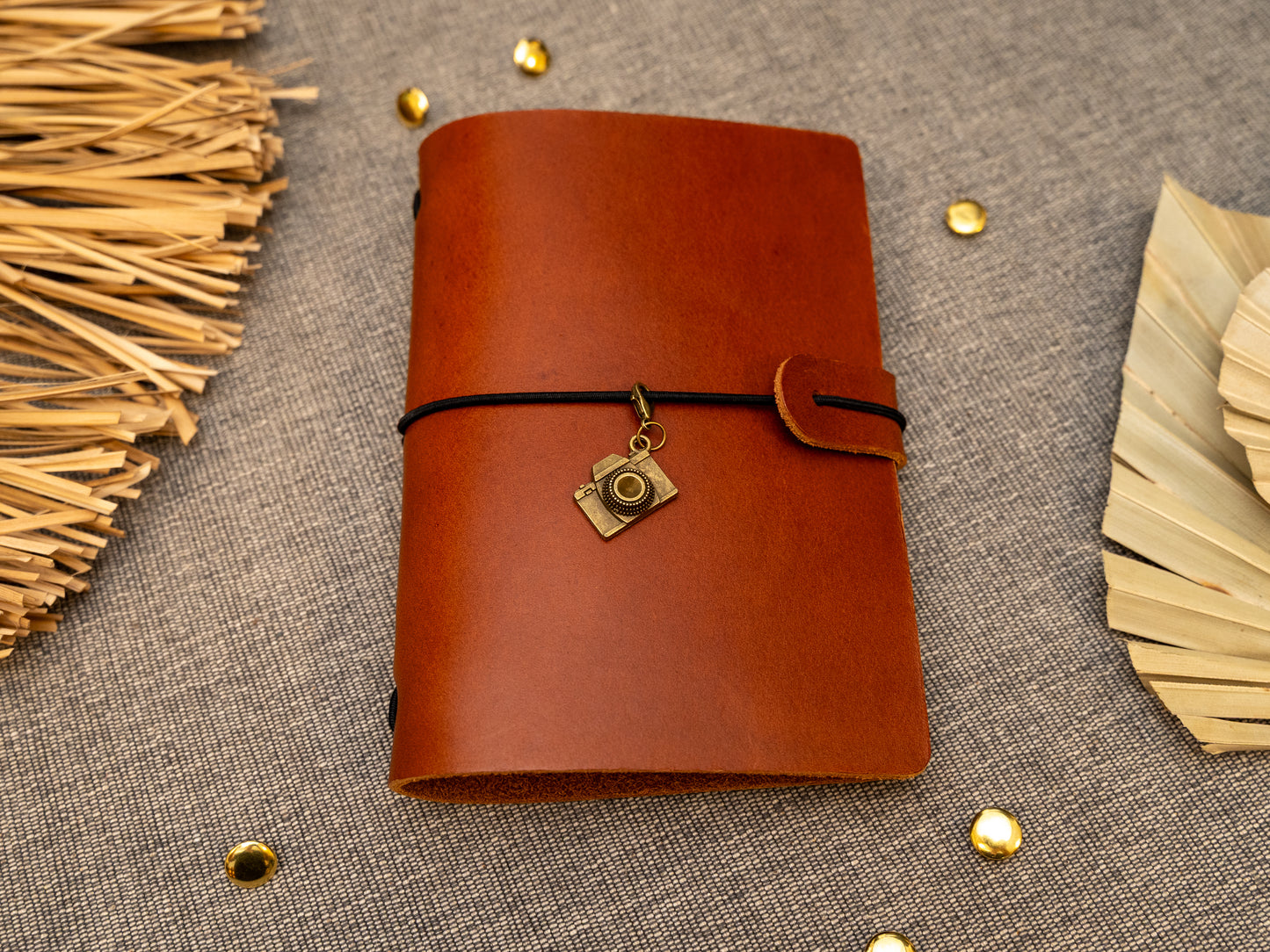A tan leather journal with a black elastic band closure and a vintage brass camera charm, displayed on a textured gray fabric background with decorative props.
