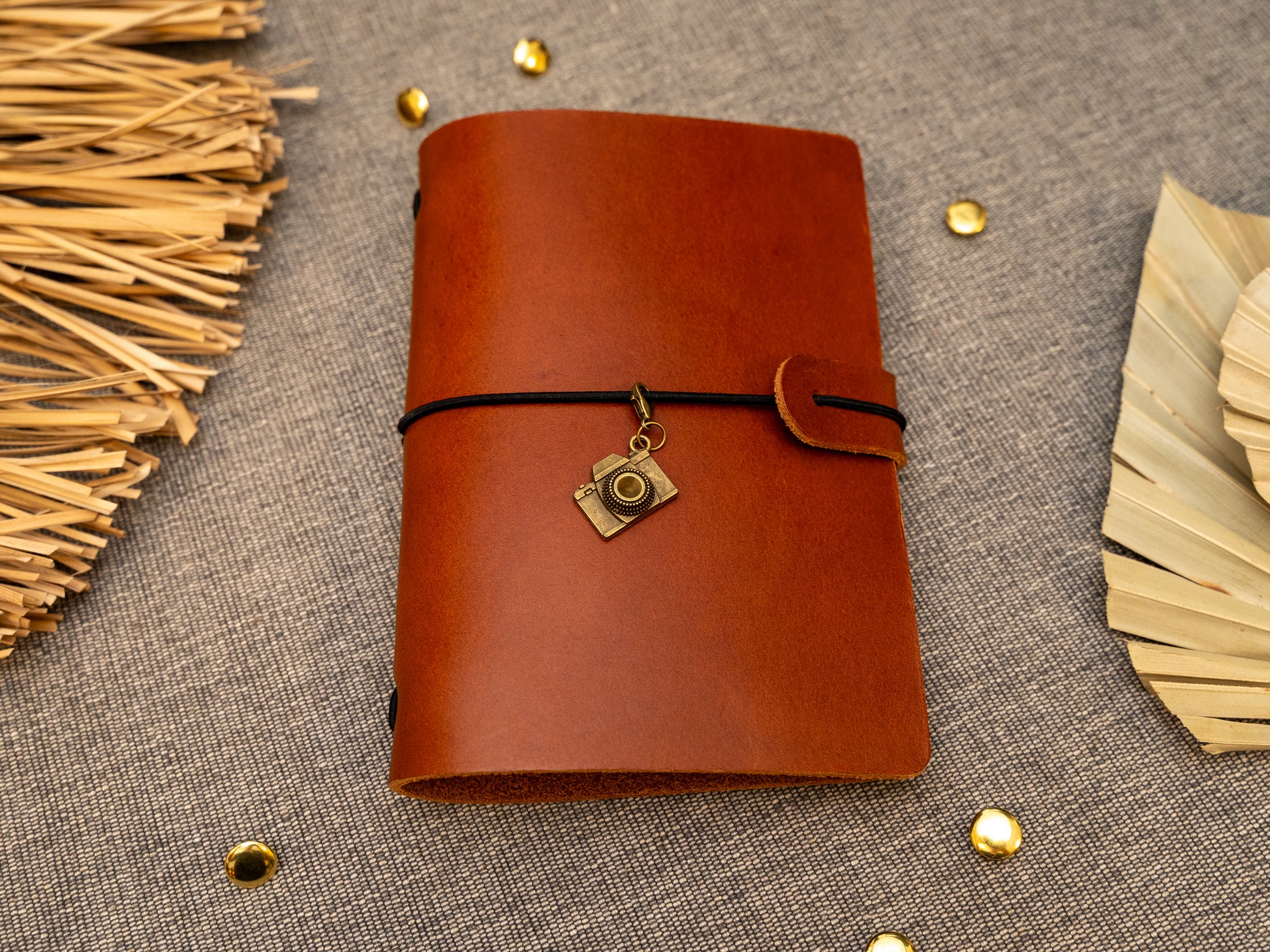A tan leather journal with a black elastic band closure and a vintage brass camera charm, displayed on a textured gray fabric background with decorative props.