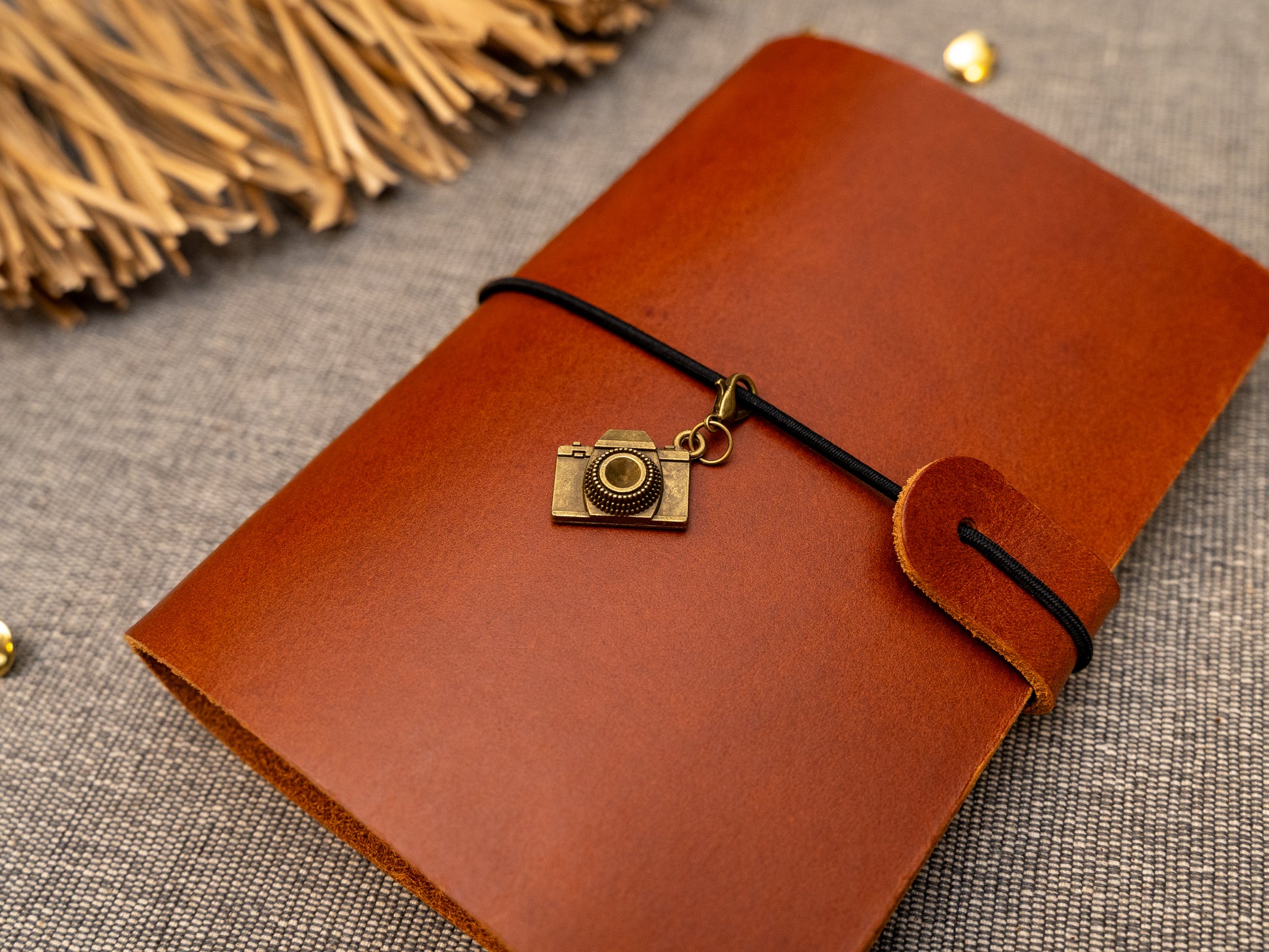 A tan leather journal with a black elastic band closure and a vintage brass camera charm, displayed on a textured gray fabric background with decorative props.