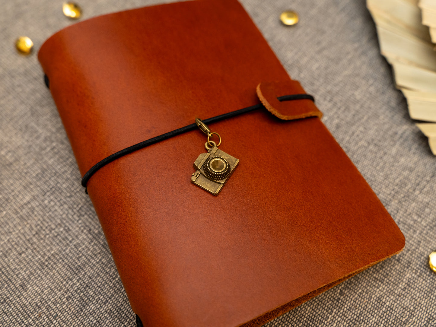 A tan leather journal with a black elastic band closure and a vintage brass camera charm, displayed on a textured gray fabric background with decorative props.