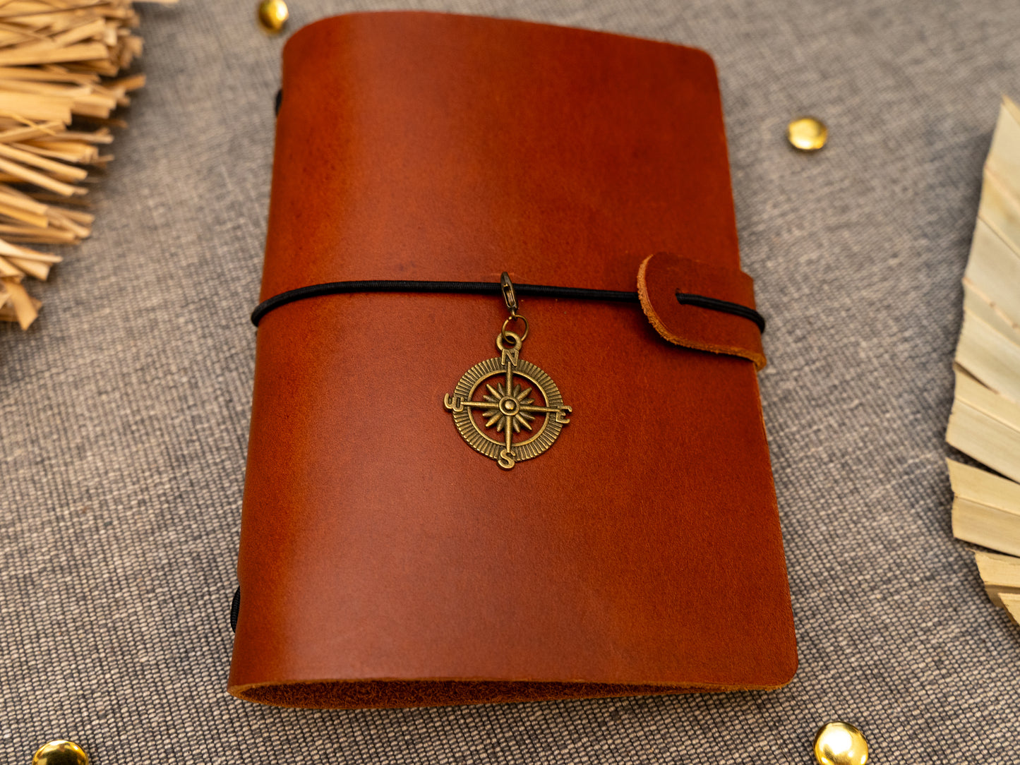 A tan leather journal with a black elastic band closure and a vintage brass compass charm, displayed on a textured gray fabric background with decorative props.