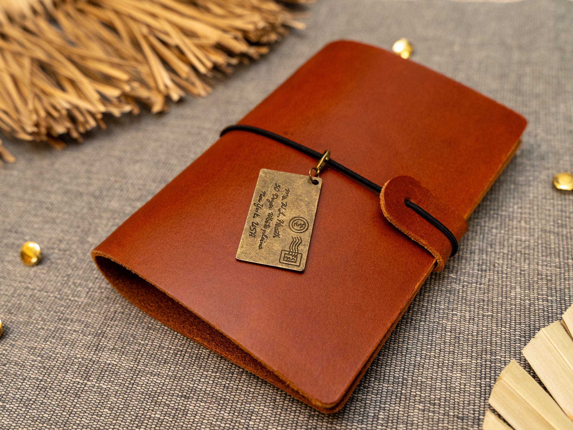 A tan leather journal with a black elastic band closure and a vintage brass envelope charm, displayed on a textured gray fabric background with decorative props.