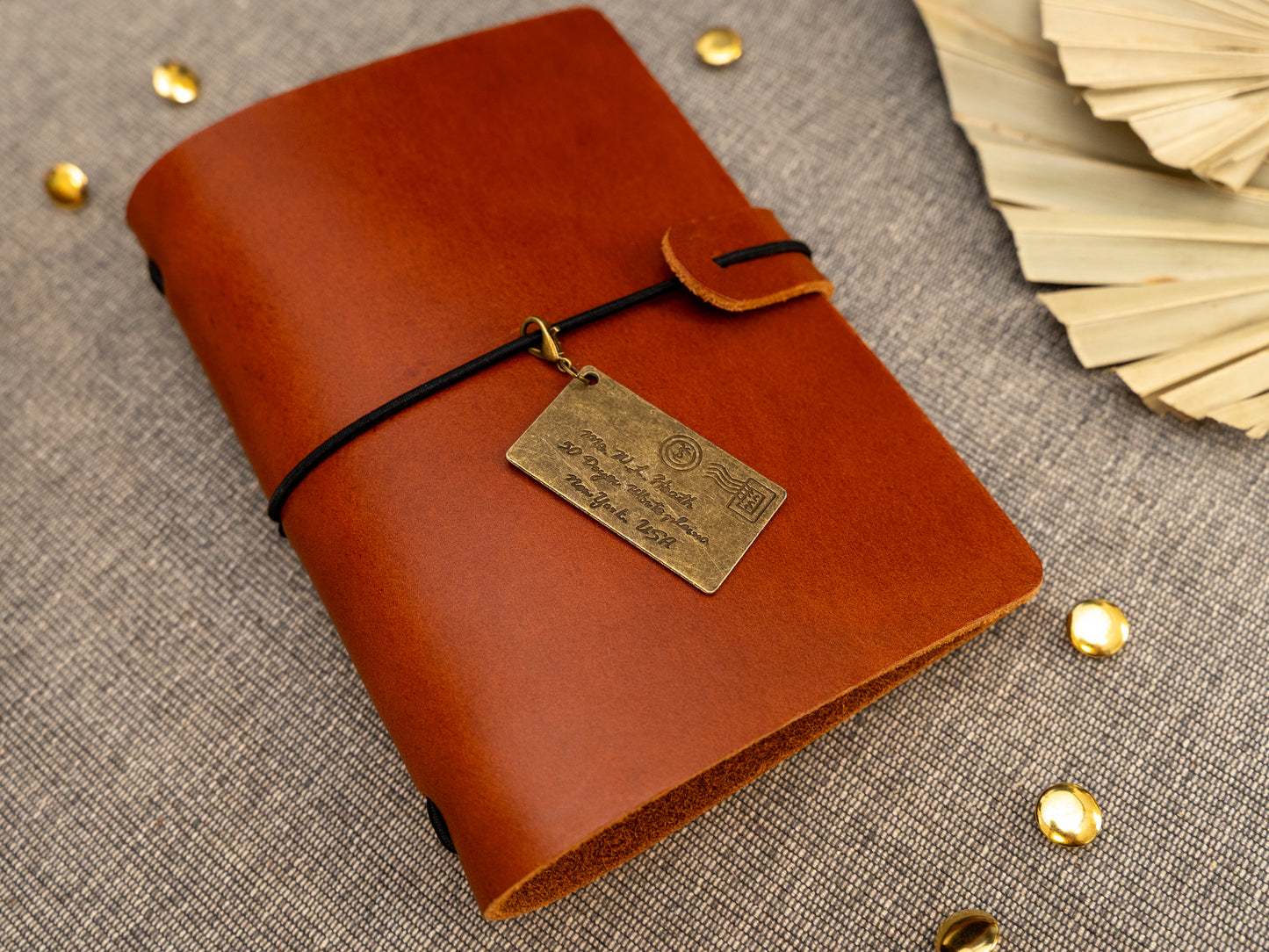 A tan leather journal with a black elastic band closure and a vintage brass envelope charm, displayed on a textured gray fabric background with decorative props.