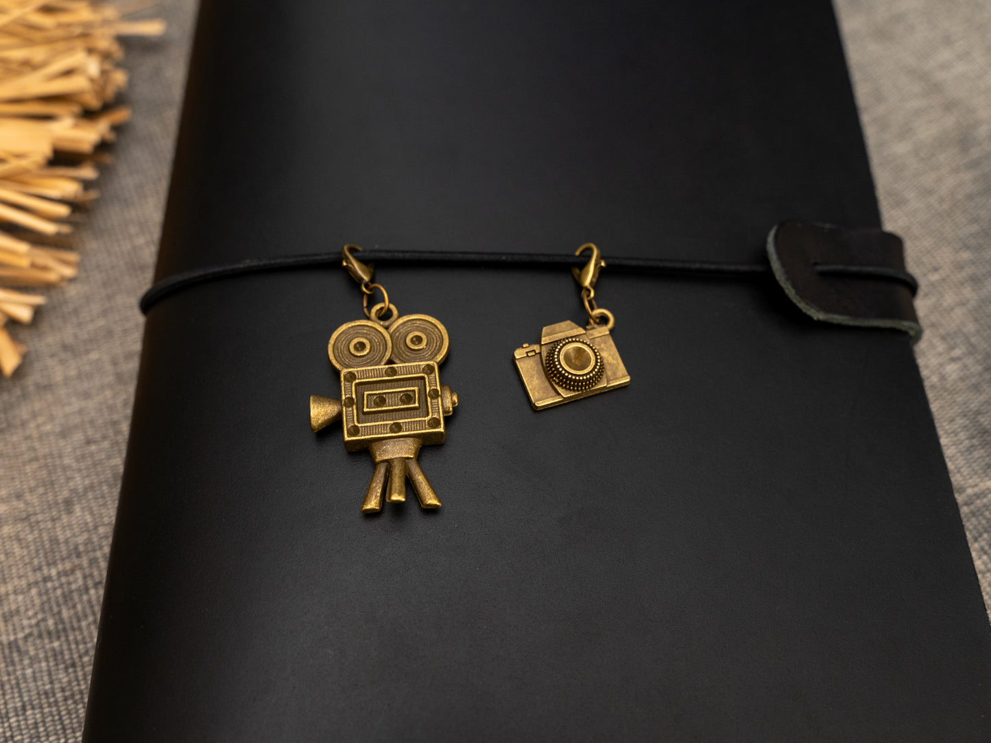 A black leather journal with a black elastic band closure and a vintage brass film camera and a camera charms, displayed clipped on to a notebook side by side
