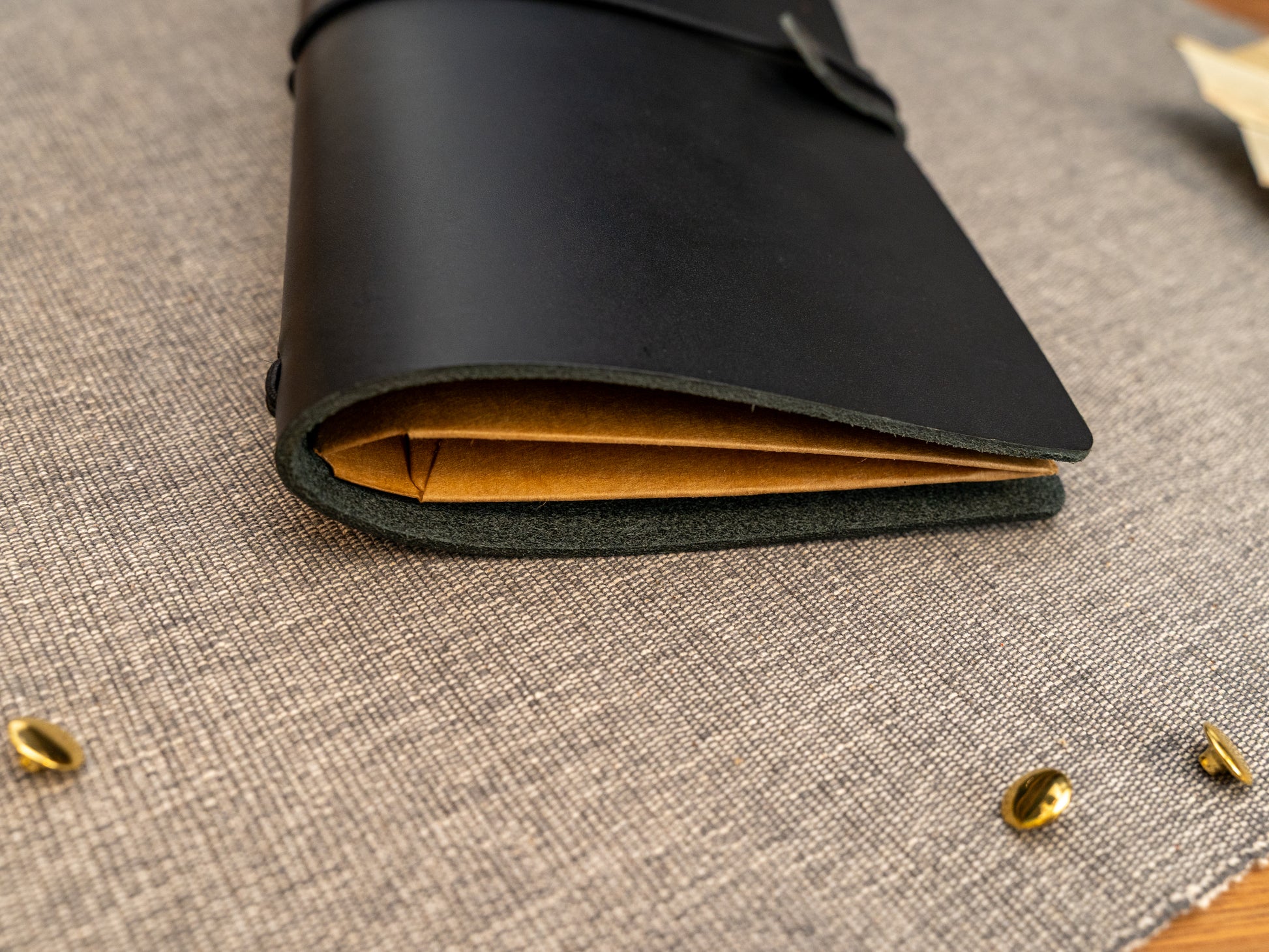 A close-up of a black leather notebook or journal cover with a Kraft card organiser insert, resting on a textured grey fabric surface with small gold metal accents