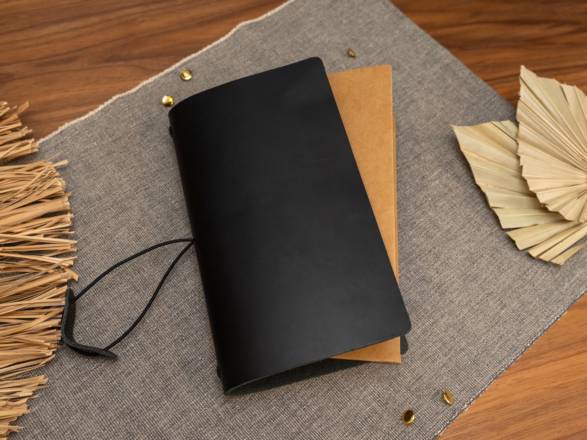A black leather journal cover with a black cord tie, shown open to reveal a Kraft card organiser, styled on a grey linen mat with dried leaves and small brass pins on a wooden surface.