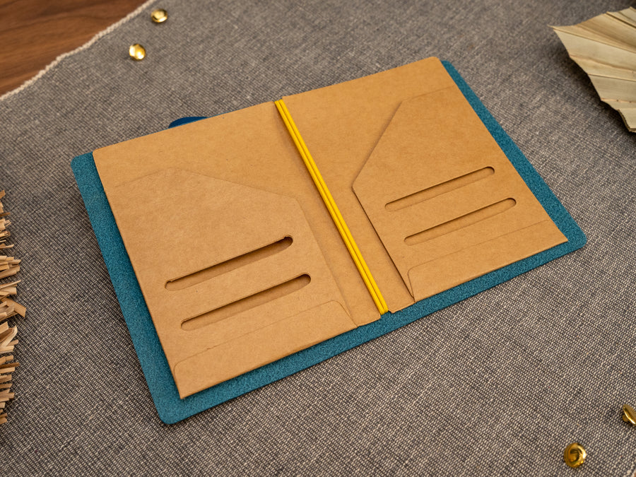An open tn passport size Kraft card document holder or organizer in a leather notebook cover, shown on a textured gray fabric surface with decorative dried leaves and small gold accents.