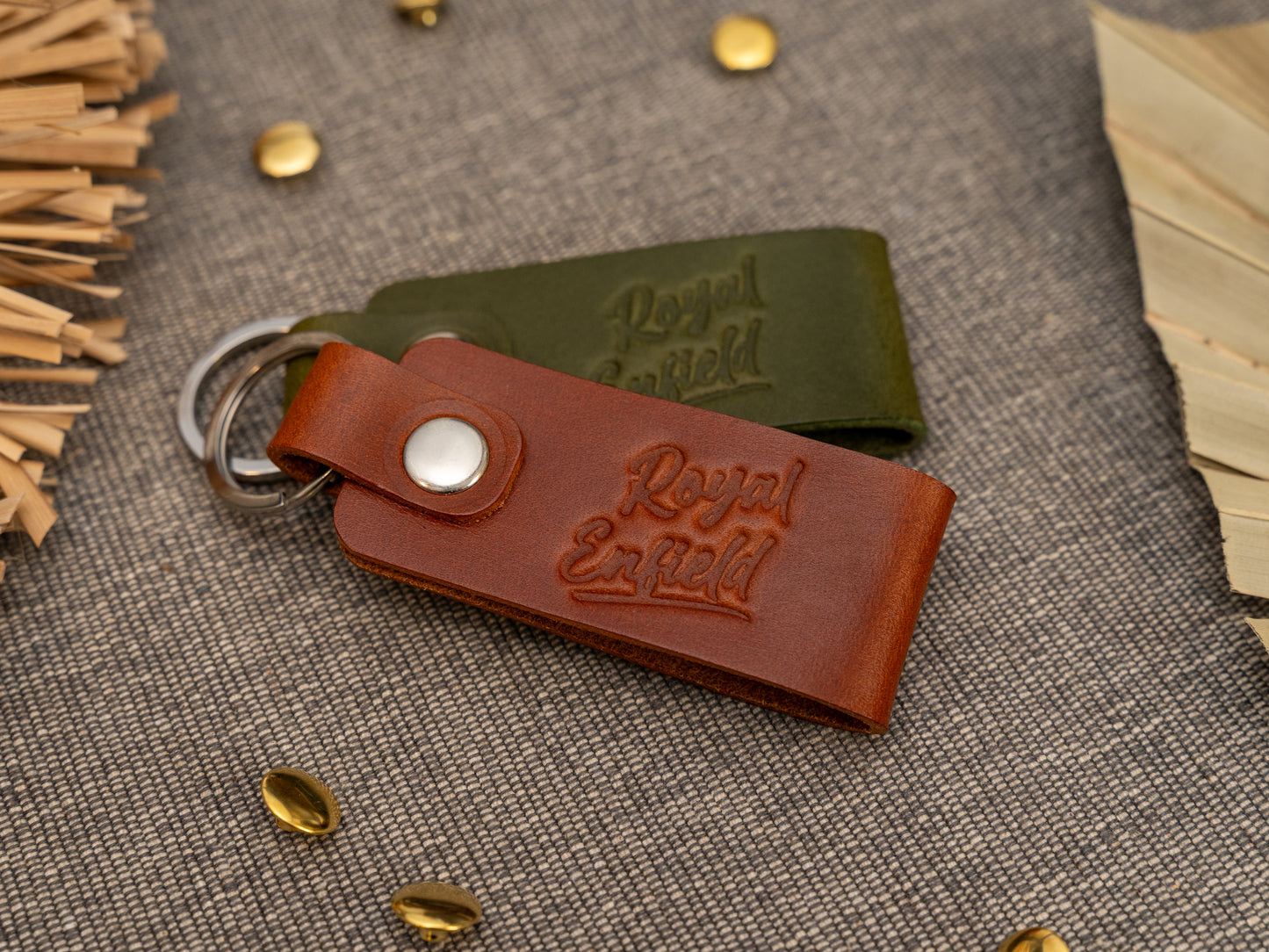 Folded tan and olive green leather keyrings/keychains with Royal Enfield in a cursive style font debossed on them. Made from full grain leather with stainless steel split ring and rivet.