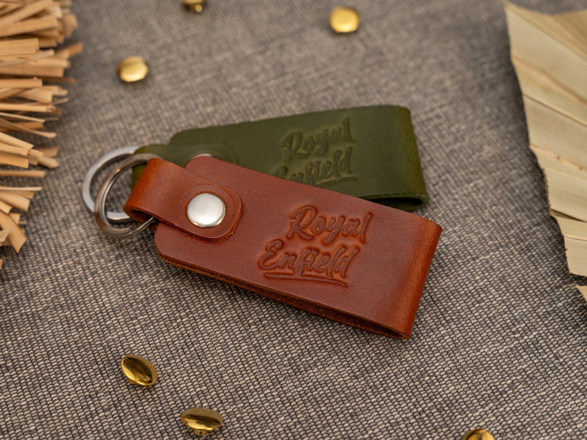 Folded tan and olive green leather keyrings/keychains with Royal Enfield in a cursive style font debossed on them. Made from full grain leather with stainless steel split ring and rivet.