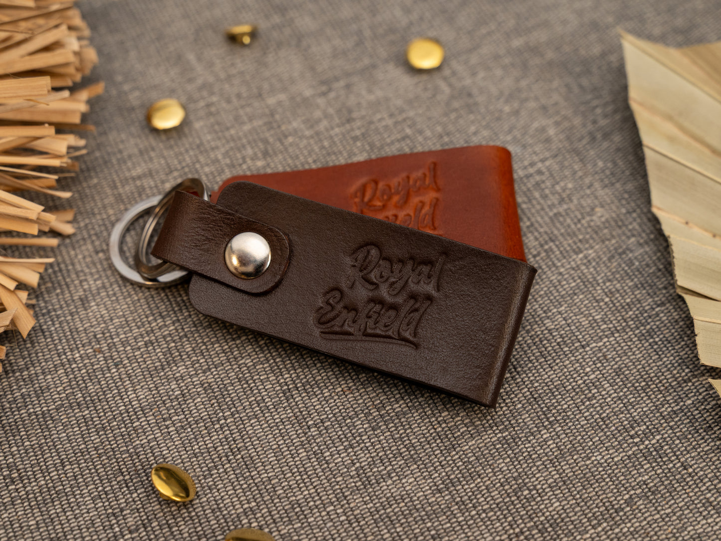 Folded brown and tan leather keyrings/keychains with Royal Enfield in a cursive style font debossed on them. Made from full grain leather with stainless steel split ring and rivet.