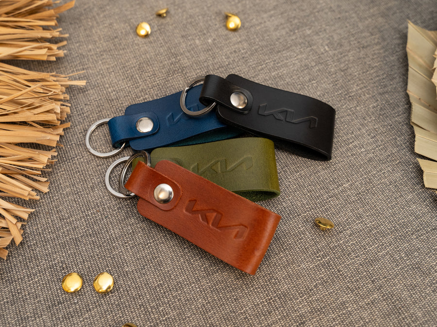 Several keyrings with a folded design in various colours such as tan, olive green, navy blue and black keyrings/keychains embossed with the Kia car logo. Stainless steel split ring and rivet.