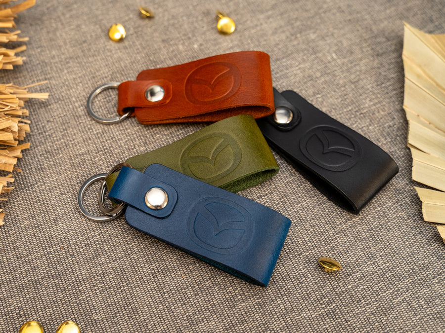 Several keyrings with a folded design in various colours such as tan, olive green, navy blue and black keyrings/keychains embossed with the Mazda car logo. Stainless steel split ring and rivet.