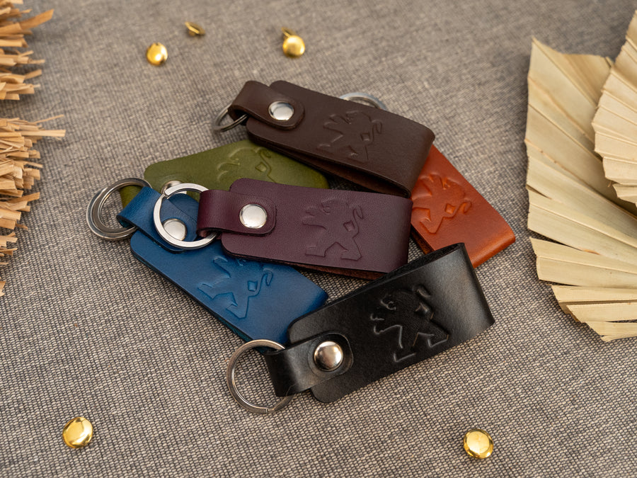 A group of folded leather keyrings in blue, tan, olive green, purple, dark brown and black leather with Peugeot car logo debossed on them. Made from Italian full grain leather