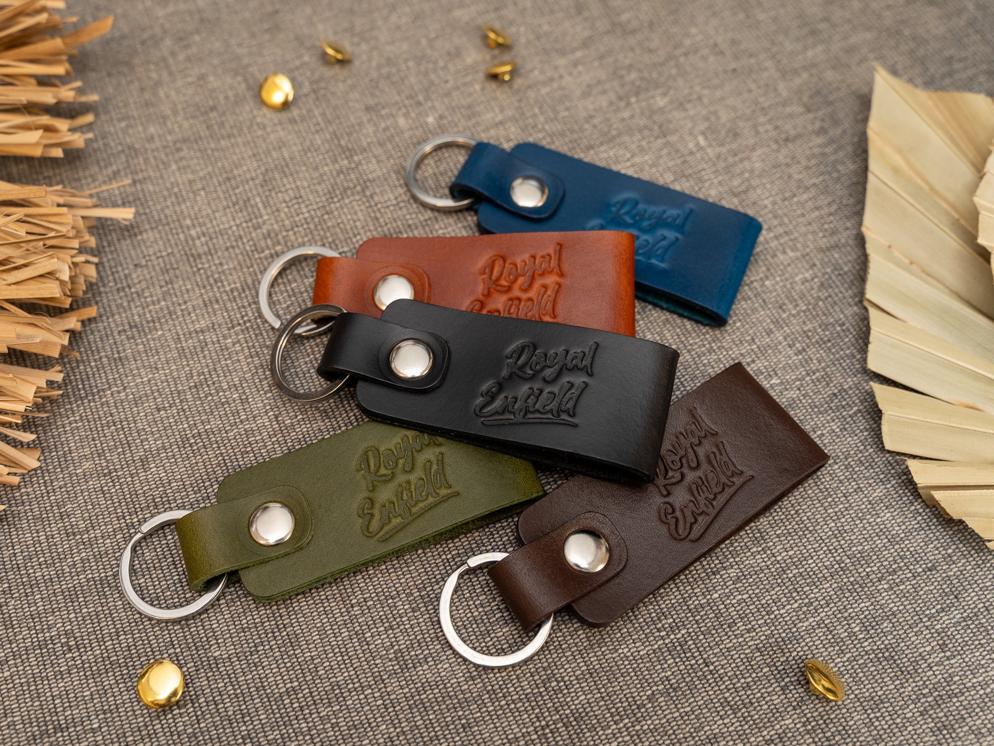 Several keyrings with a folded design in various colours such as tan, olive green, navy blue, dark brown and black keyrings/keychains embossed with Royal Enfield in a cursive style font. Stainless steel split ring and rivet.