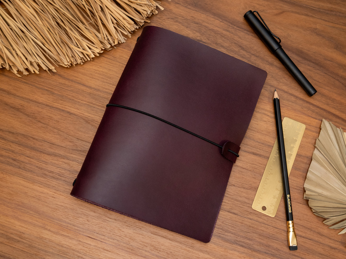 A dark purple leather journal with a black elastic closure strap, displayed on a wooden desk alongside stationery items including a gold pen, pencils, a ruler, and a woven straw mat.